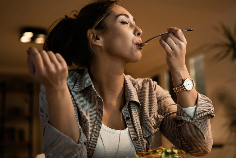 Low angle view of young woman eating salad with her eyes closed while using computer at night at home.