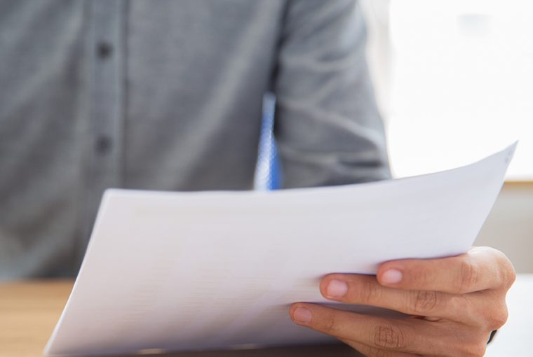 Unrecognizable businessman sitting at table, holding mobile phone and paper, reading document and texting message